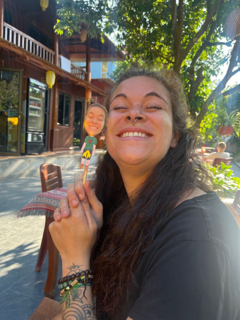 A participant smiling next to her own paper puppet self under sunlit trees.